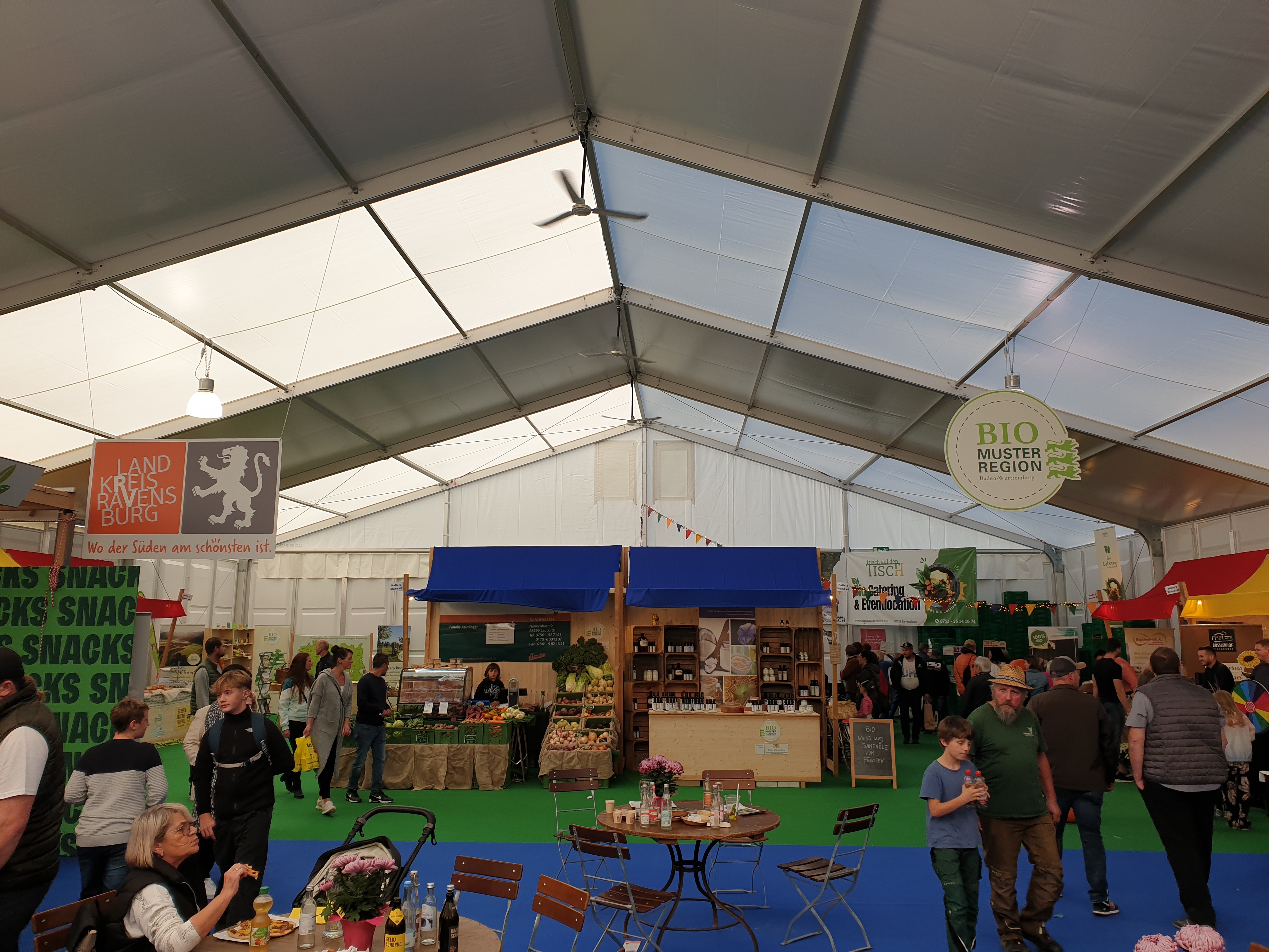 Stände des Gemeinschaftsstandes sind auf dem grünen Teppich zu sehen. Von der Zeltdecke hängen die Logos der Bio-Musterregion und des Landkreises Ravensburg.